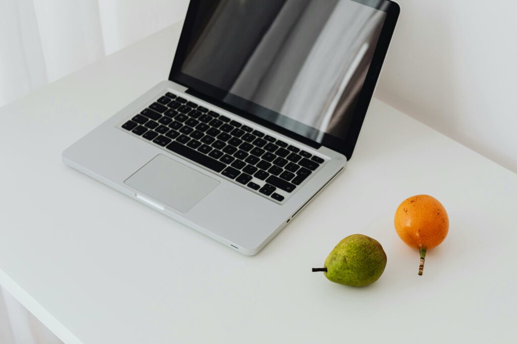 Gray and black laptop on a white desk. An orange and pear are off to the right side adding some color. Picture is from Pexels.com number 6958733