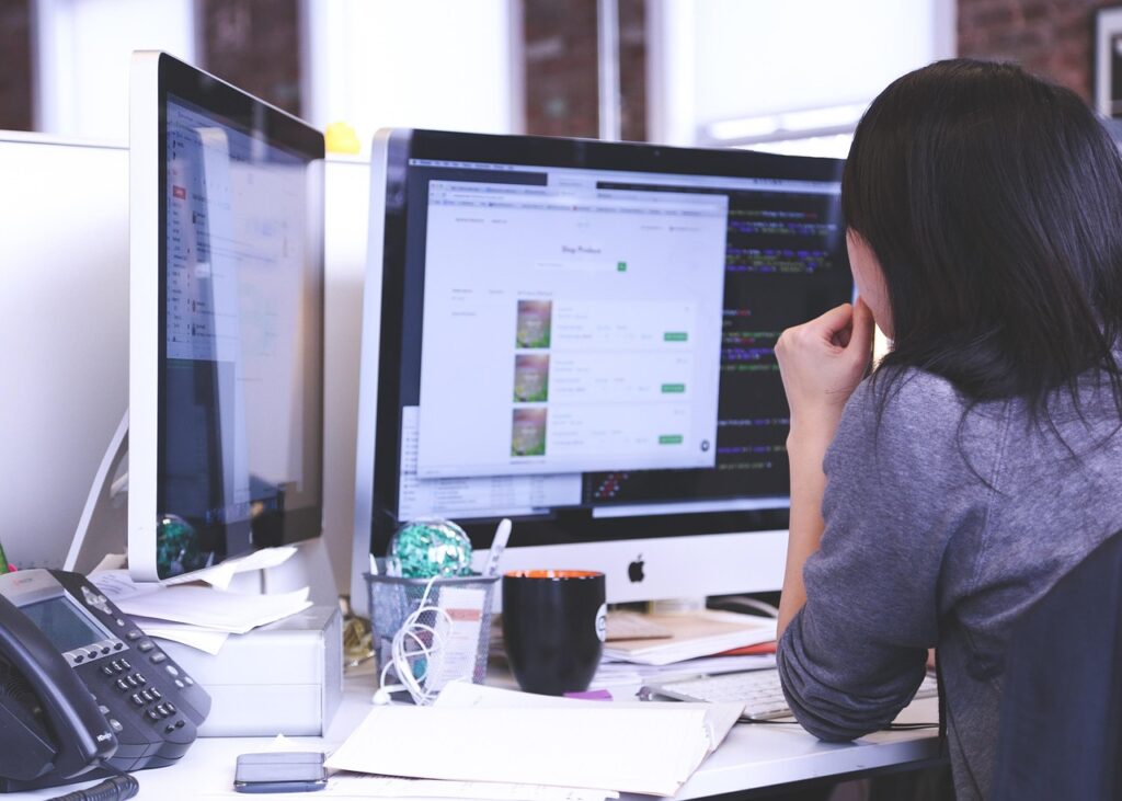 A business person in a cubicle looking at data on two different computer monitors.