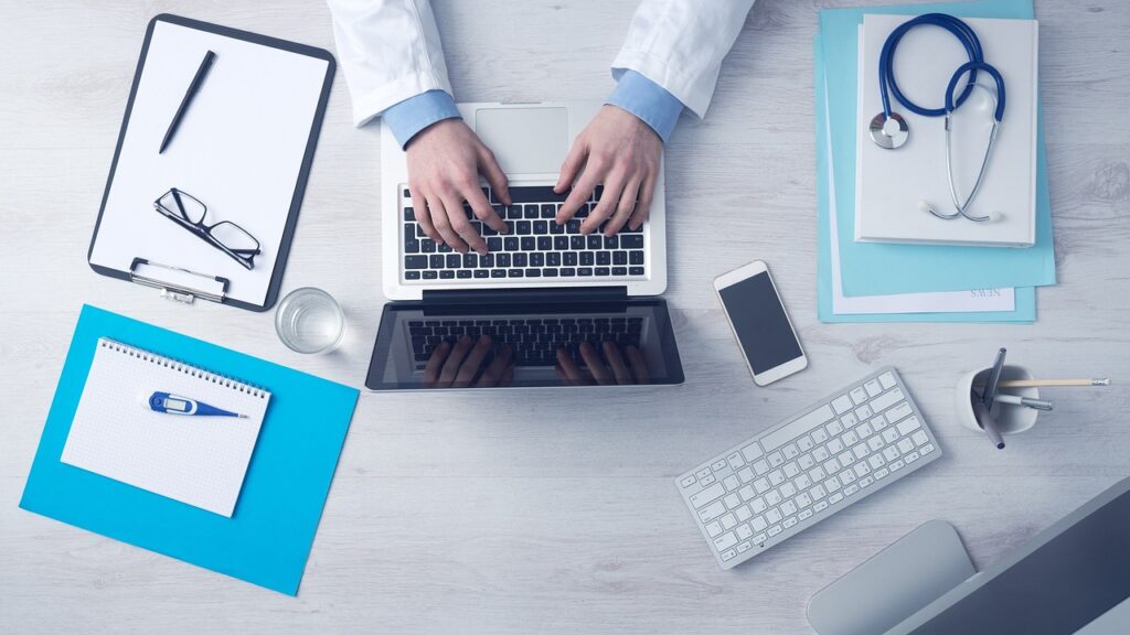 Layout view of a desk with a clipboard and stethoscope and a doctors hands typing on a laptop in the middle.